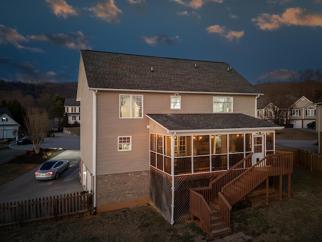 back of house at dusk featuring brick siding, a shingled roof, stairway, a sunroom, and fence