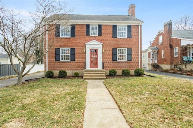 colonial home featuring brick siding, a front lawn, a chimney, and fence