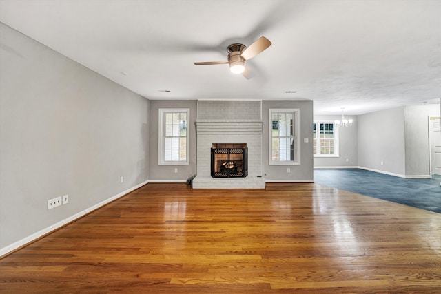 unfurnished living room featuring ceiling fan with notable chandelier, a brick fireplace, wood finished floors, and baseboards