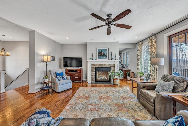 living room with a stone fireplace, recessed lighting, ceiling fan with notable chandelier, wood finished floors, and baseboards