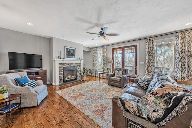 living room featuring ceiling fan, a textured ceiling, a stone fireplace, wood finished floors, and baseboards