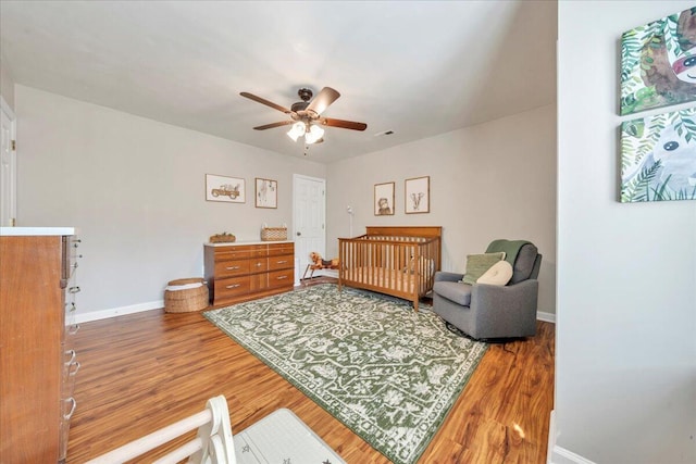 bedroom featuring a nursery area, ceiling fan, baseboards, and wood finished floors