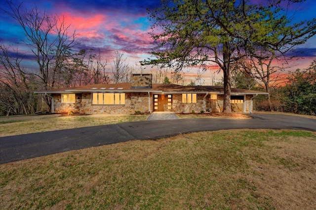 mid-century modern home featuring an attached garage, a lawn, a chimney, stone siding, and driveway