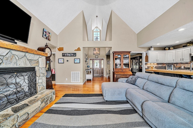 living area featuring a textured ceiling, high vaulted ceiling, a stone fireplace, visible vents, and light wood-style floors