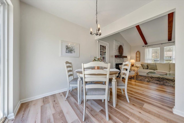 dining area with baseboards, lofted ceiling with beams, light wood-style flooring, an inviting chandelier, and a stone fireplace