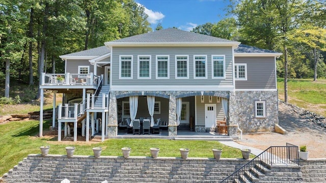 rear view of property with a patio, a shingled roof, stairs, stone siding, and a lawn