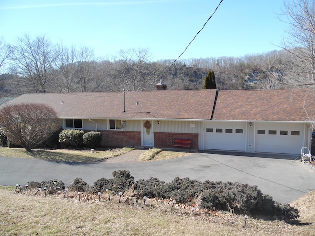 single story home featuring a garage, brick siding, driveway, roof with shingles, and a chimney