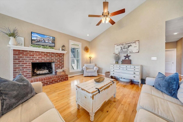 living room with high vaulted ceiling, light wood finished floors, a brick fireplace, and a ceiling fan