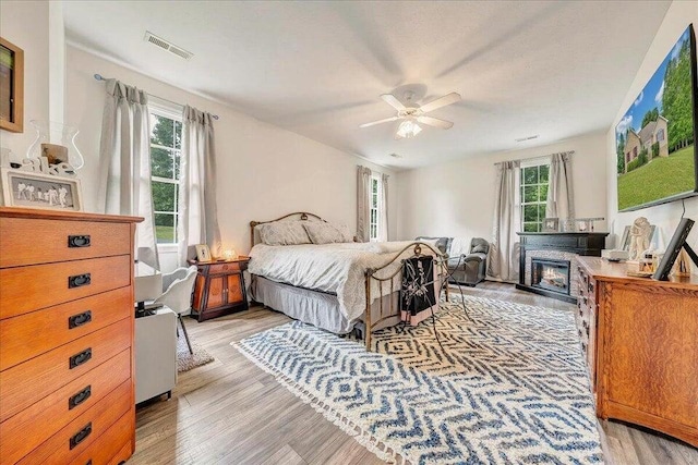 bedroom featuring light wood-style floors, visible vents, a ceiling fan, and a glass covered fireplace