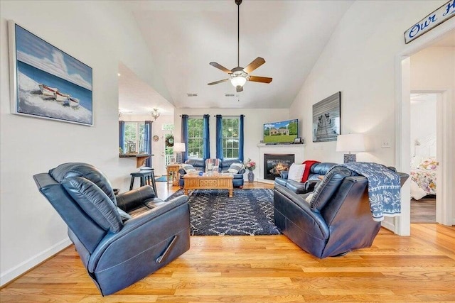 living room featuring a ceiling fan, baseboards, vaulted ceiling, light wood finished floors, and a glass covered fireplace