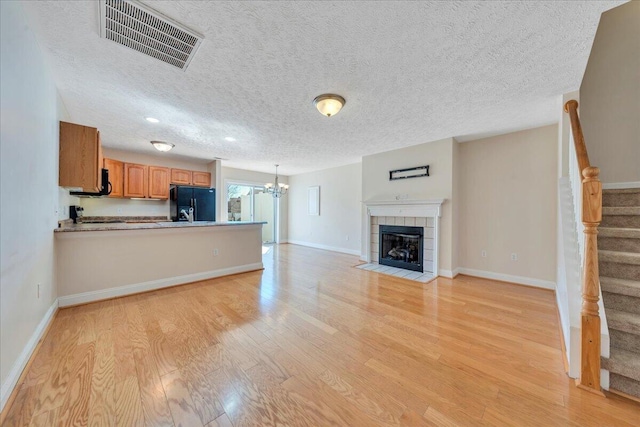 unfurnished living room with visible vents, stairs, light wood-style floors, a tiled fireplace, and a chandelier