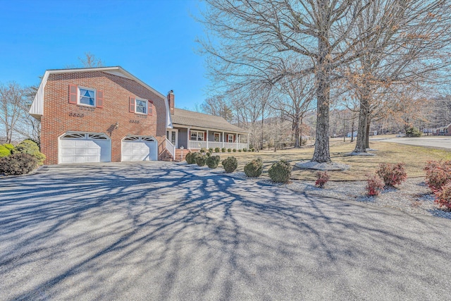 view of front of home featuring brick siding, a chimney, covered porch, a garage, and driveway