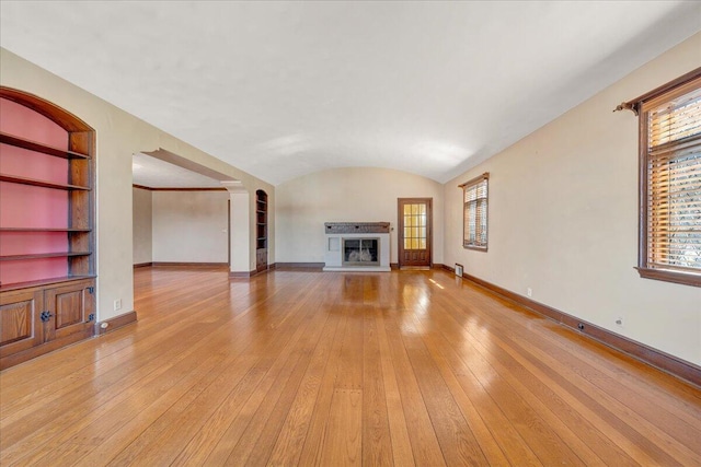 unfurnished living room with built in shelves, light wood-style floors, a glass covered fireplace, vaulted ceiling, and baseboards