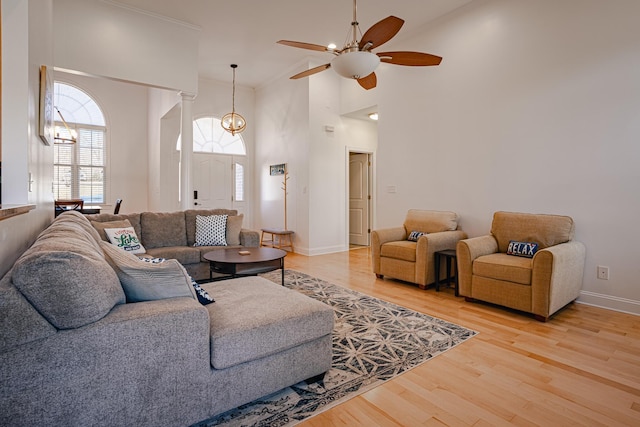 living area with baseboards, a towering ceiling, light wood-type flooring, ornate columns, and ceiling fan with notable chandelier