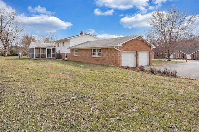 view of property exterior featuring driveway, a garage, a lawn, a sunroom, and brick siding