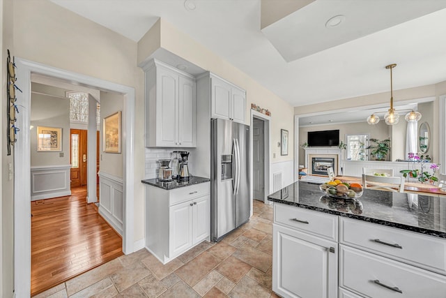 kitchen with stainless steel fridge, white cabinets, dark stone counters, light stone finish floors, and wainscoting