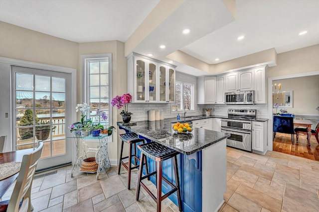 kitchen with appliances with stainless steel finishes, dark stone countertops, a peninsula, glass insert cabinets, and white cabinets