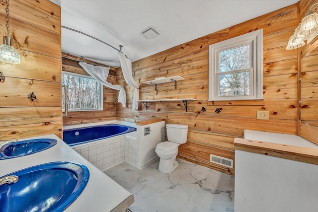 full bathroom featuring tiled bath, visible vents, wood walls, toilet, and a sink