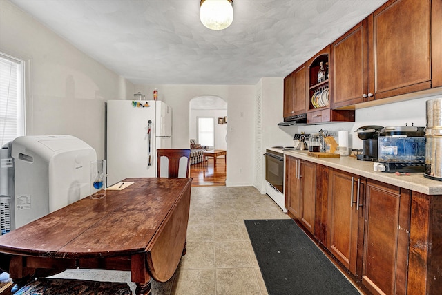 kitchen with brown cabinets, range with electric cooktop, open shelves, freestanding refrigerator, and arched walkways