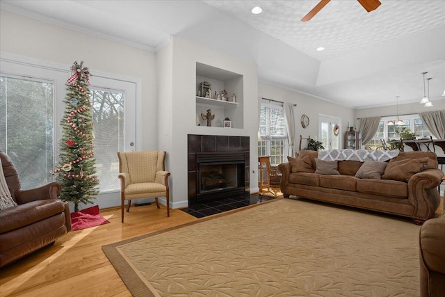 living room with built in shelves, a ceiling fan, a tiled fireplace, wood finished floors, and crown molding