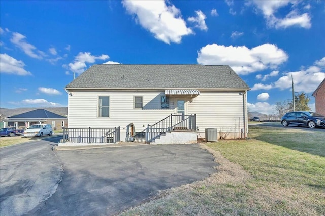 back of house with aphalt driveway, central AC unit, a shingled roof, and a yard