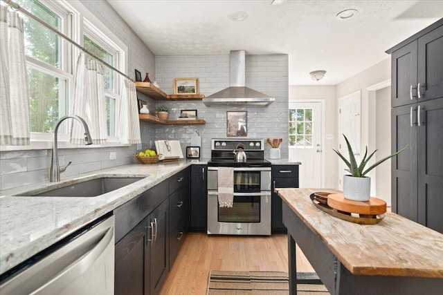 kitchen with light wood finished floors, a sink, appliances with stainless steel finishes, wall chimney exhaust hood, and backsplash