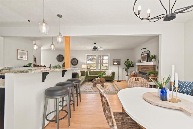 dining area with ceiling fan with notable chandelier, a brick fireplace, and light wood finished floors