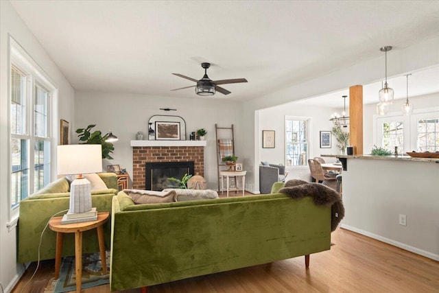 living room featuring ceiling fan with notable chandelier, a brick fireplace, baseboards, and wood finished floors