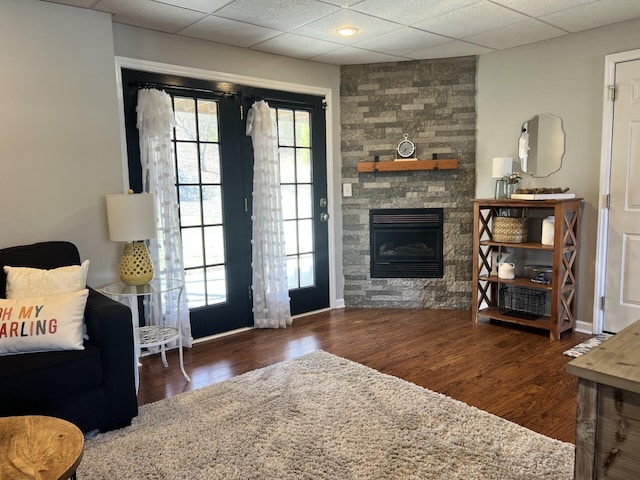 living room featuring a stone fireplace, wood finished floors, baseboards, and a drop ceiling