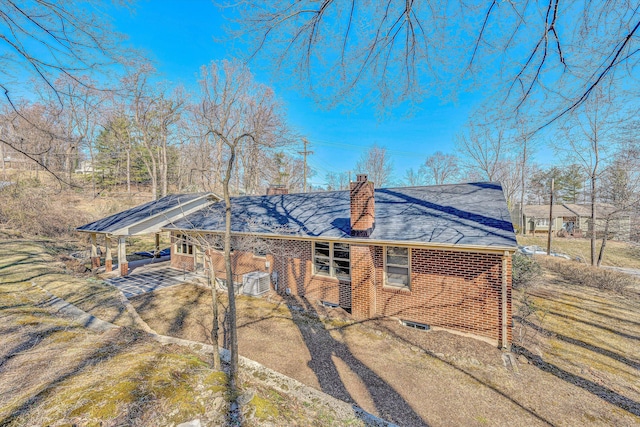 view of home's exterior with brick siding, a shingled roof, a chimney, a carport, and driveway