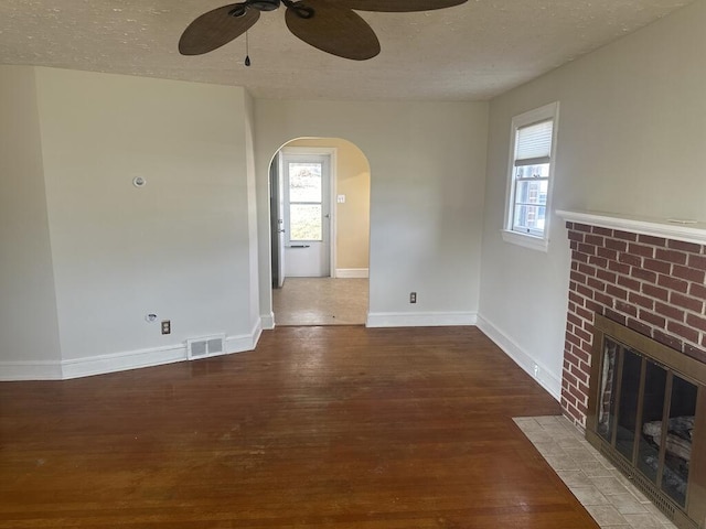 unfurnished living room with visible vents, wood finished floors, arched walkways, a brick fireplace, and ceiling fan