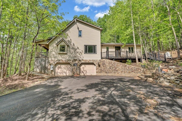 rustic home with an attached garage, a wooded view, a wooden deck, and aphalt driveway