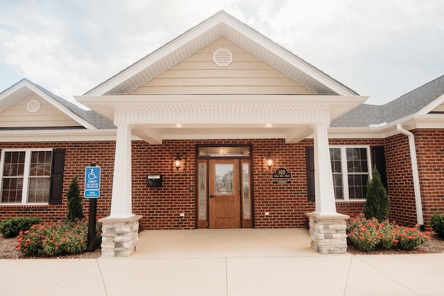 doorway to property featuring brick siding, covered porch, and roof with shingles