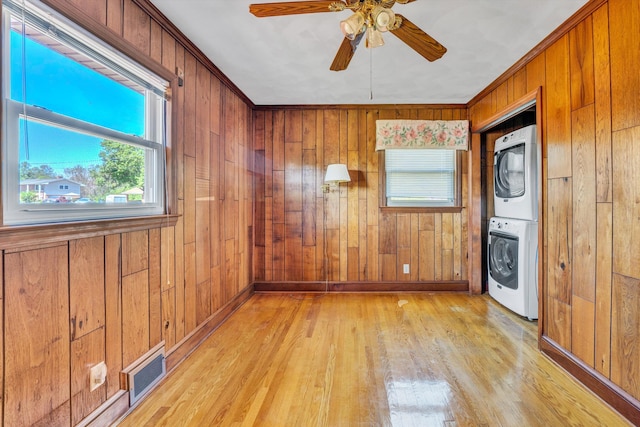 empty room featuring wooden walls, stacked washer / drying machine, ceiling fan, light wood-style floors, and visible vents