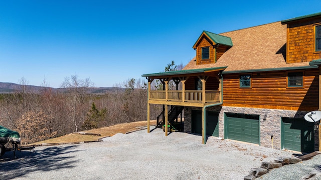 rear view of house with driveway, stone siding, an attached garage, and roof with shingles