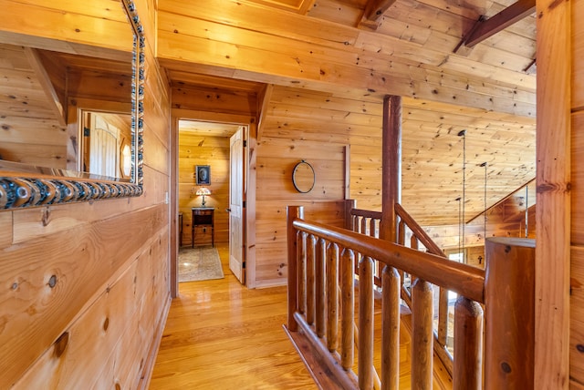 hallway with an upstairs landing, wooden walls, light wood finished floors, beam ceiling, and wooden ceiling