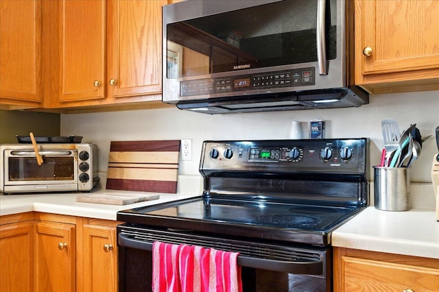 kitchen featuring light countertops, black / electric stove, stainless steel microwave, brown cabinetry, and a toaster