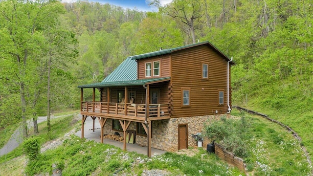 back of house with a wooded view, stone siding, a deck, metal roof, and log exterior