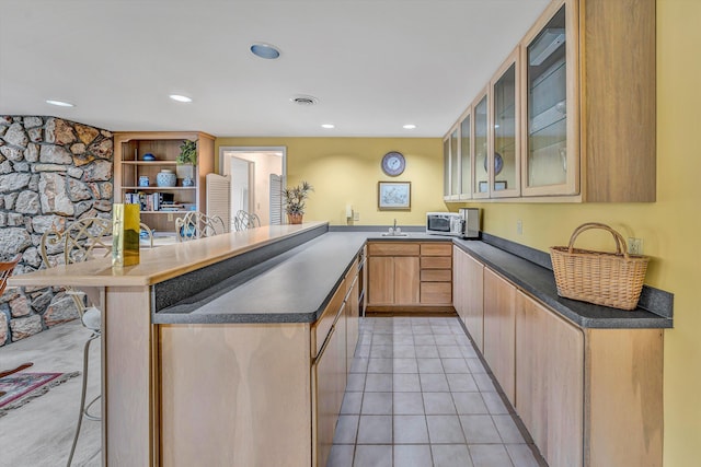 kitchen featuring a kitchen breakfast bar, dark countertops, a sink, light tile patterned flooring, and glass insert cabinets