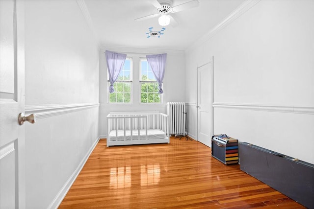 unfurnished bedroom featuring radiator, crown molding, wood finished floors, a crib, and a ceiling fan