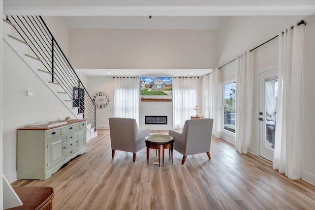 sitting room with baseboards, light wood finished floors, a towering ceiling, stairs, and a glass covered fireplace