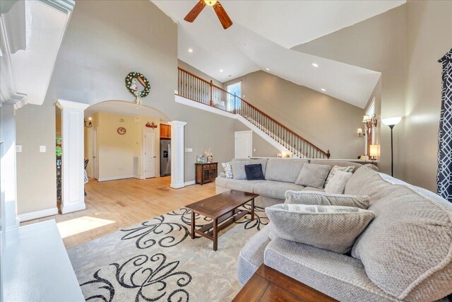 living room featuring high vaulted ceiling, stairway, ceiling fan, light wood finished floors, and decorative columns