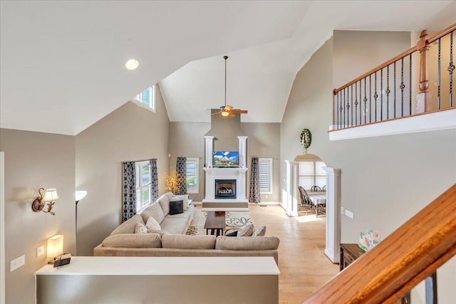 living room with a fireplace with raised hearth, ornate columns, light wood-style floors, high vaulted ceiling, and ceiling fan