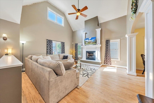 living room with light wood-type flooring, a fireplace with raised hearth, ornate columns, a ceiling fan, and high vaulted ceiling
