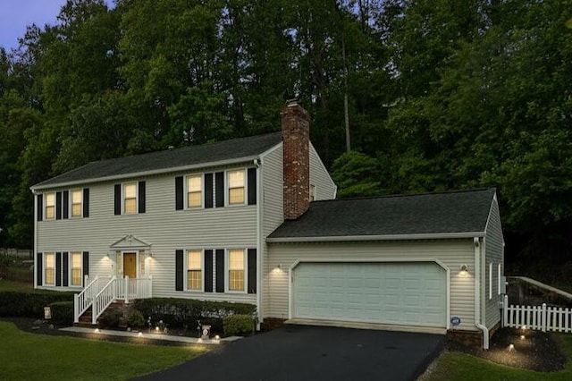 colonial-style house featuring driveway, an attached garage, a chimney, and view of scattered trees
