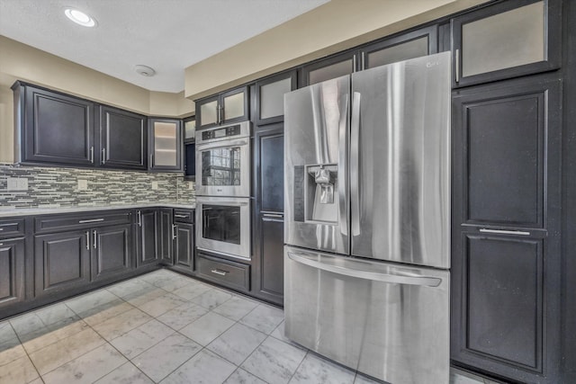 kitchen featuring stainless steel appliances, backsplash, glass insert cabinets, marble finish floor, and light countertops