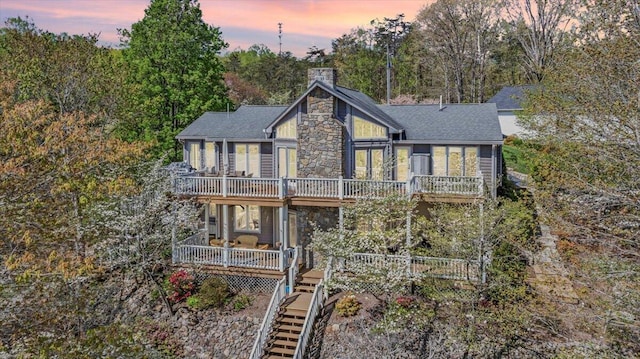 back of house at dusk with a chimney, stone siding, a deck, roof with shingles, and stairs