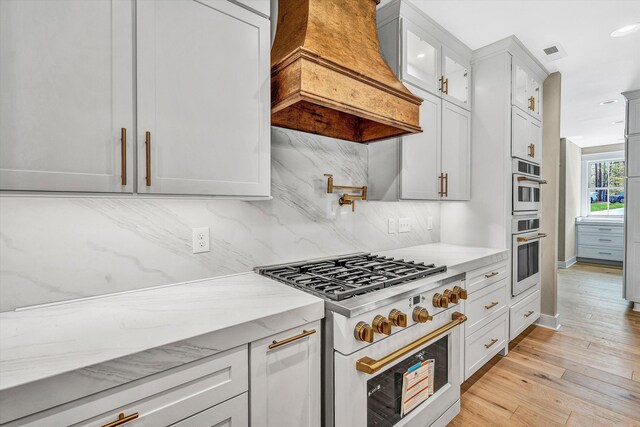 kitchen featuring light wood finished floors, decorative backsplash, gas stove, custom range hood, and light stone countertops