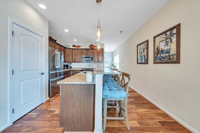 kitchen featuring stainless steel appliances, a kitchen island with sink, wood finished floors, a kitchen bar, and light stone countertops
