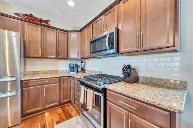 kitchen with stainless steel appliances, wood finished floors, decorative backsplash, light stone counters, and brown cabinets